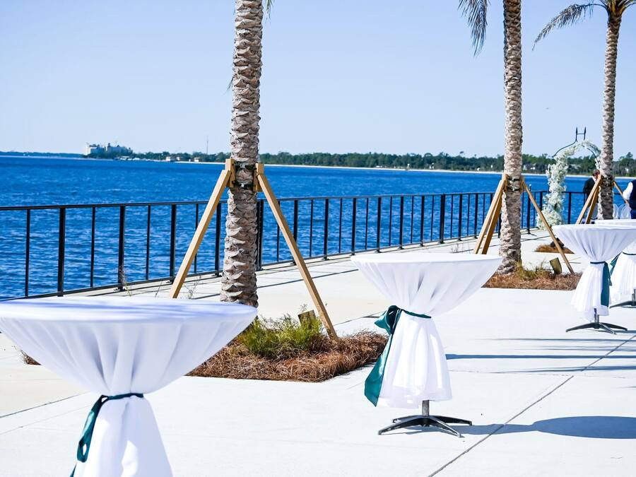Outdoor tables with white tablecloths and teal ribbons, set against a backdrop of a coastal view with palm trees and a railing.