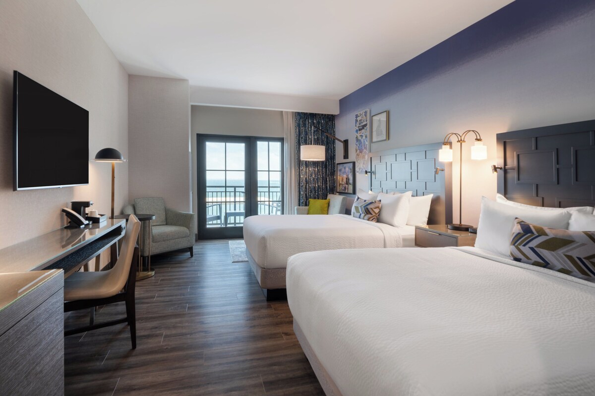 A hotel room with two beds, a desk, a chair, a TV, and a balcony with a view.