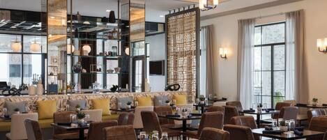 A modern dining area with high ceilings, chandeliers, and a mix of seating including tables and sofas.