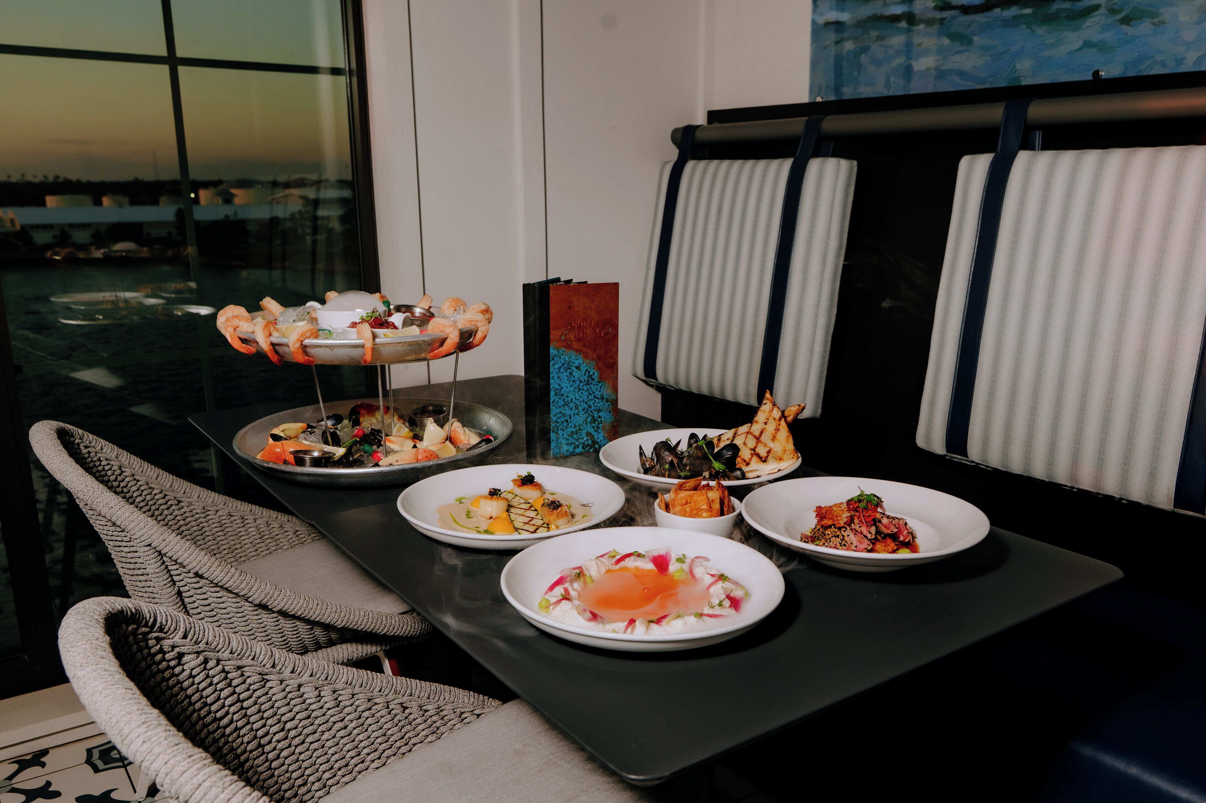 A dining table with various seafood dishes, a woven chair, and a view of the city through large windows.