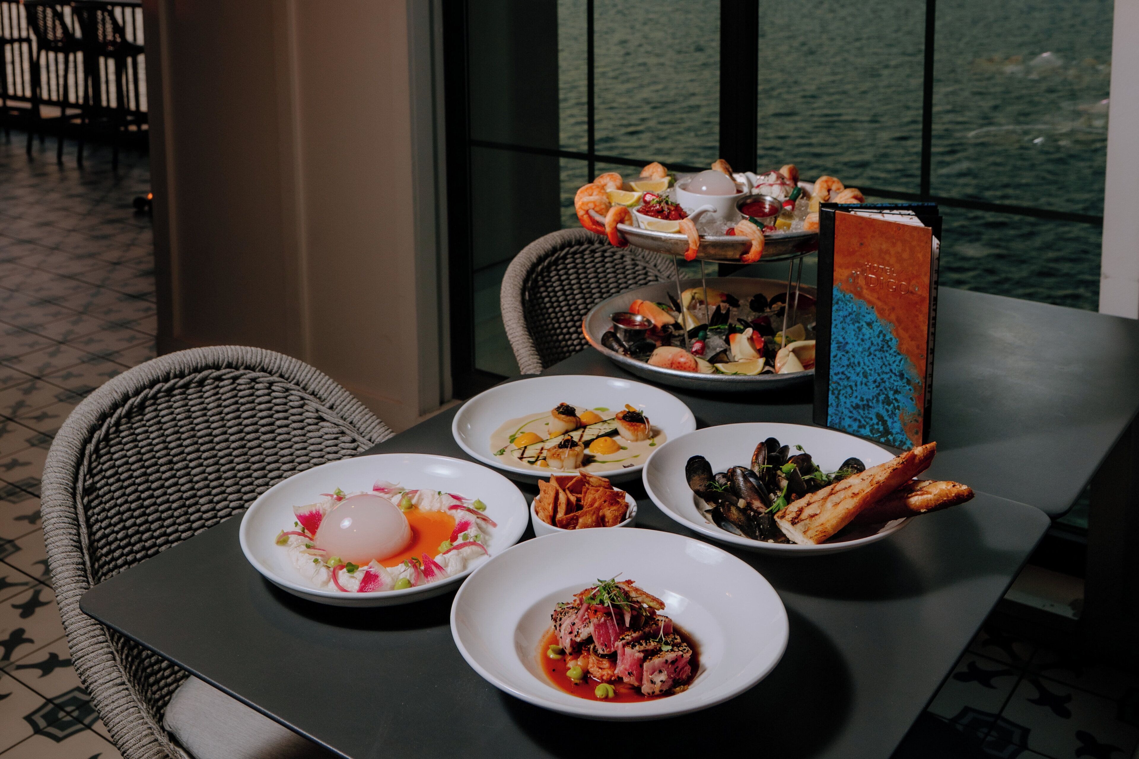 A dining table with various seafood dishes, a wicker chair, and a view of the water through large windows.