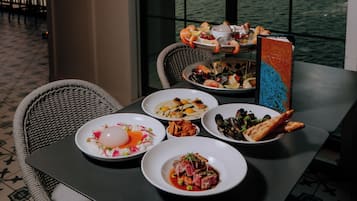 A dining table with various seafood dishes, a wicker chair, and a view of the water through large windows.