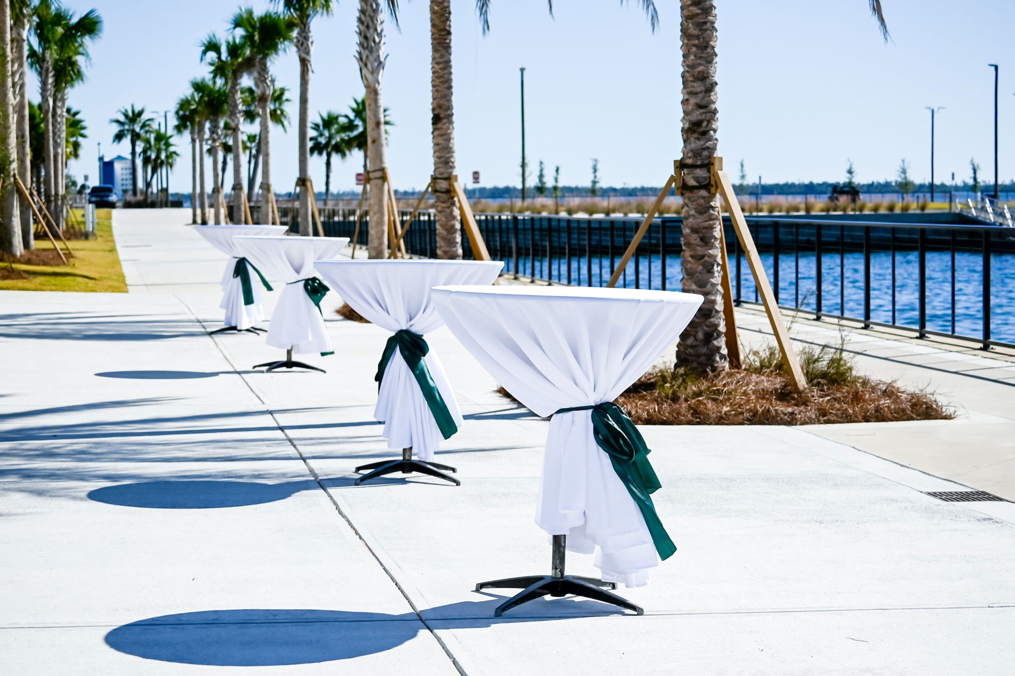 Un paseo marítimo con mesas preparadas para un evento, palmeras y una masa de agua al fondo.