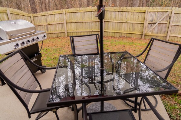 A glass-top table with four chairs, a barbecue grill, and a wooden fence in the background. - Beautiful home away from home (Raleigh)