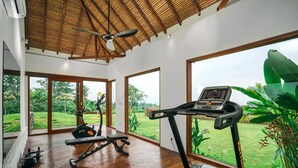 Ein geräumiger Fitnessraum mit Laufband, Ergometer und Gewichten. Große Fenster bieten Blick auf einen üppigen Garten.