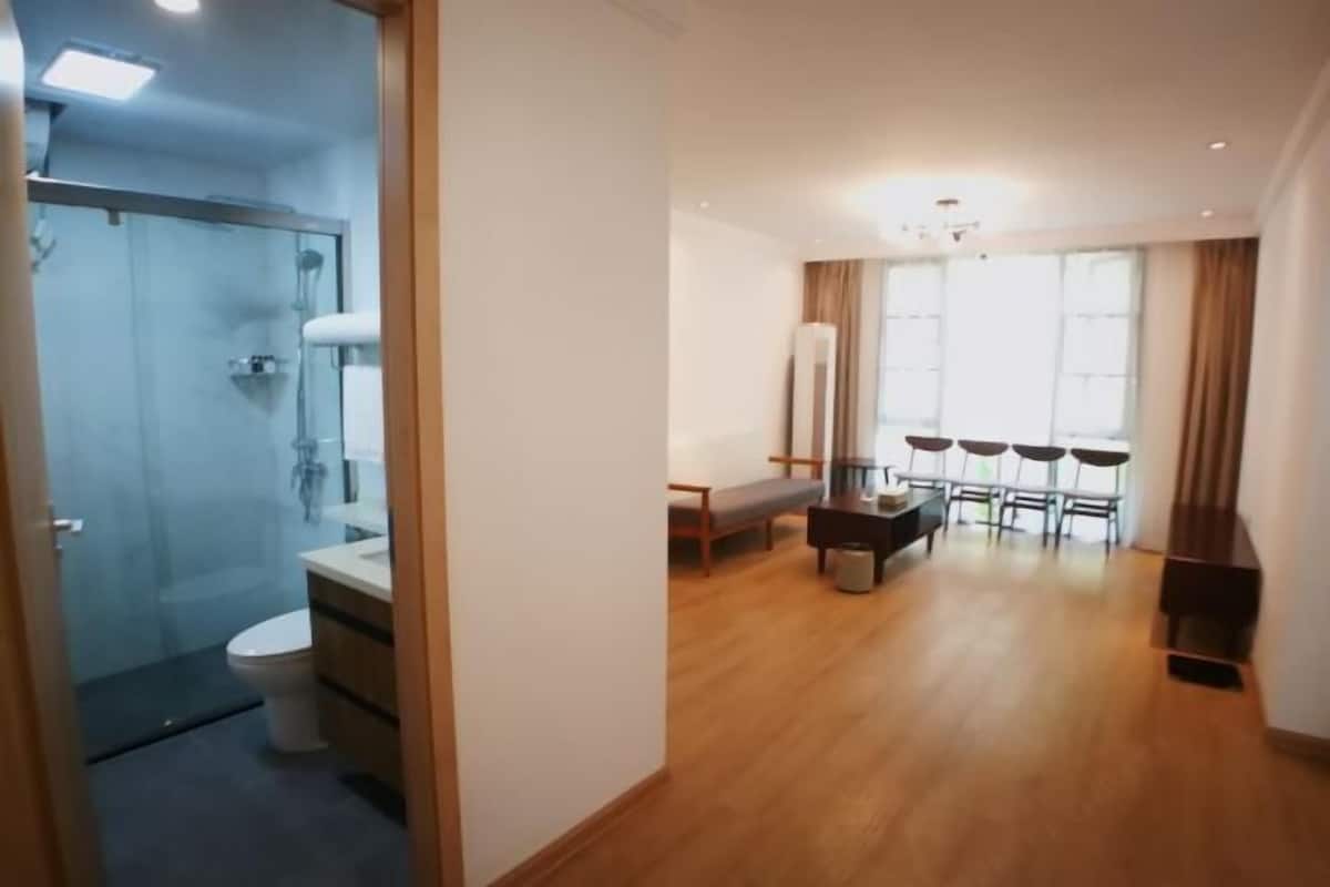 A room with a wooden floor, a bed, a bedside table, a window with curtains, and a bathroom with a toilet and shower.