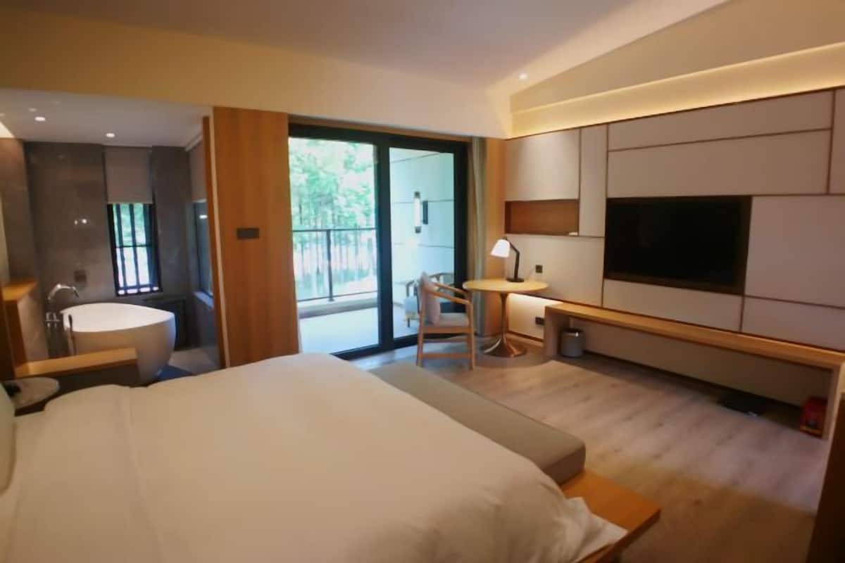 A modern hotel room with a large bed, a desk, a chair, a TV, and a balcony with a view of greenery.
