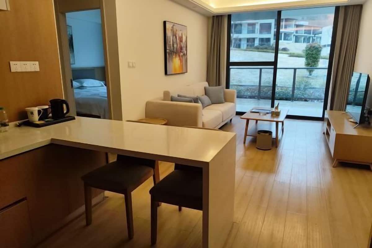 A modern hotel room with a kitchenette, a sofa, a dining table, and a large window offering a view of the outside.