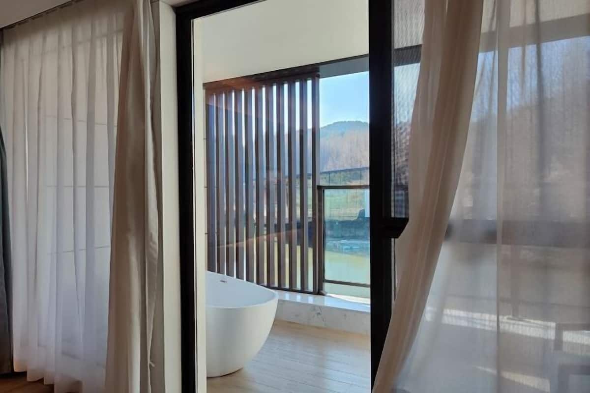 A modern bathroom with a large bathtub, a wooden floor, and a view of mountains through a window with vertical blinds.