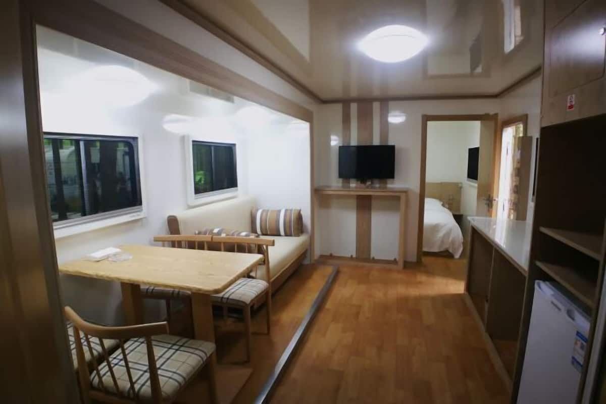 A compact room with a wooden dining table and chairs, a sofa, a television on a stand, and a refrigerator.