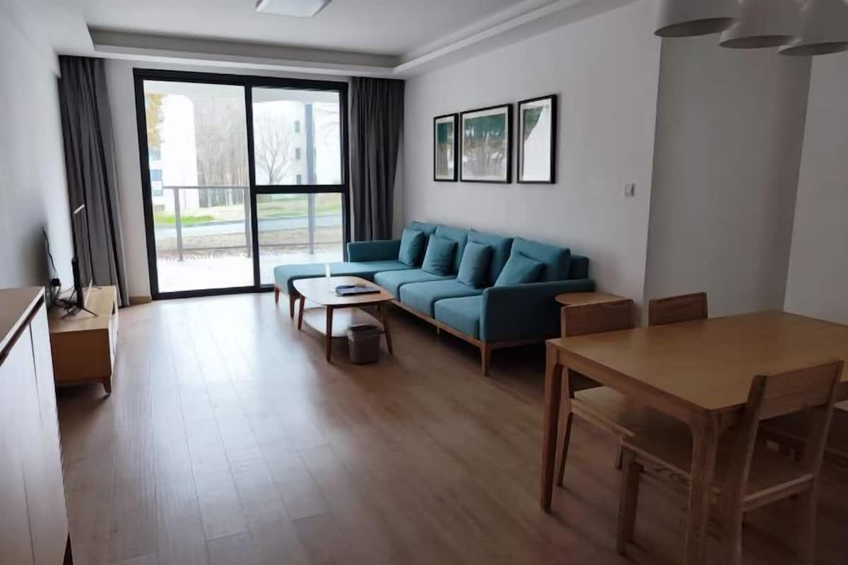 A modern living room with a sofa, a wooden dining table, and a large sliding glass door leading to an outdoor area.