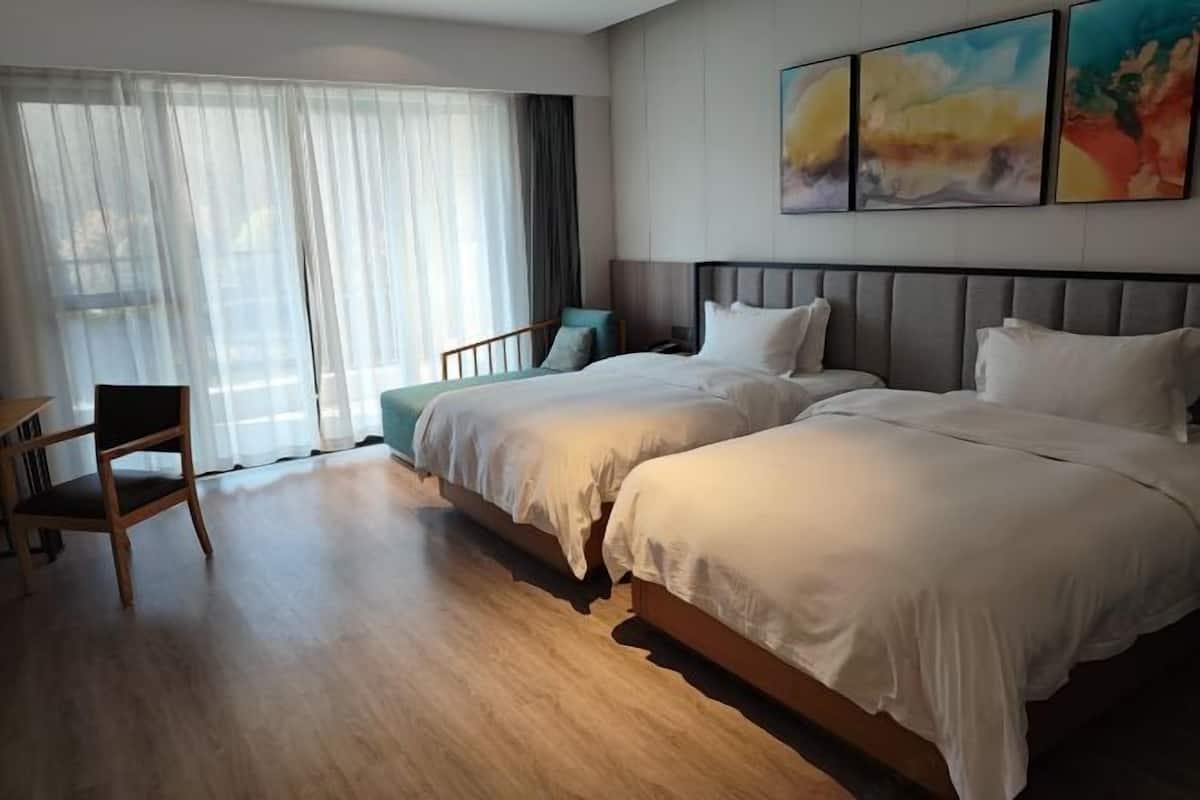 A hotel room with two beds, a desk, a chair, and a large window with sheer curtains.