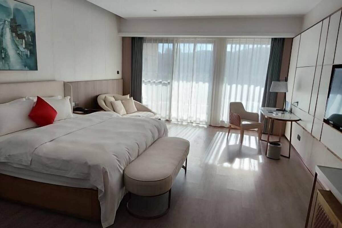 A hotel room with a large bed, a small table, a chair, a TV, and a painting on the wall.