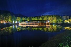 Un hôtel situé au bord de la rivière, dont l’image se reflète dans l’eau, avec un quai et une colline boisée en arrière-plan.