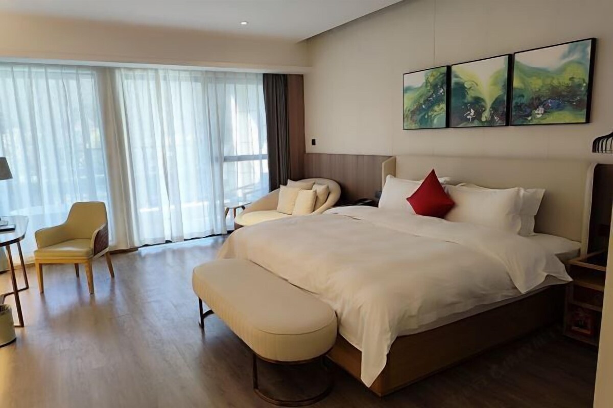 Hotellihuoneessa on suuri sänky, penkki, tuoli ja työpöytä. Seinällä on kolme taulua ja ikkunassa verhot.