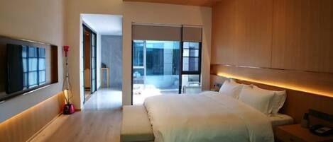 A modern bedroom with a large bed, a bedside table, a TV mounted on the wall, and a sliding glass door leading to a balcony.