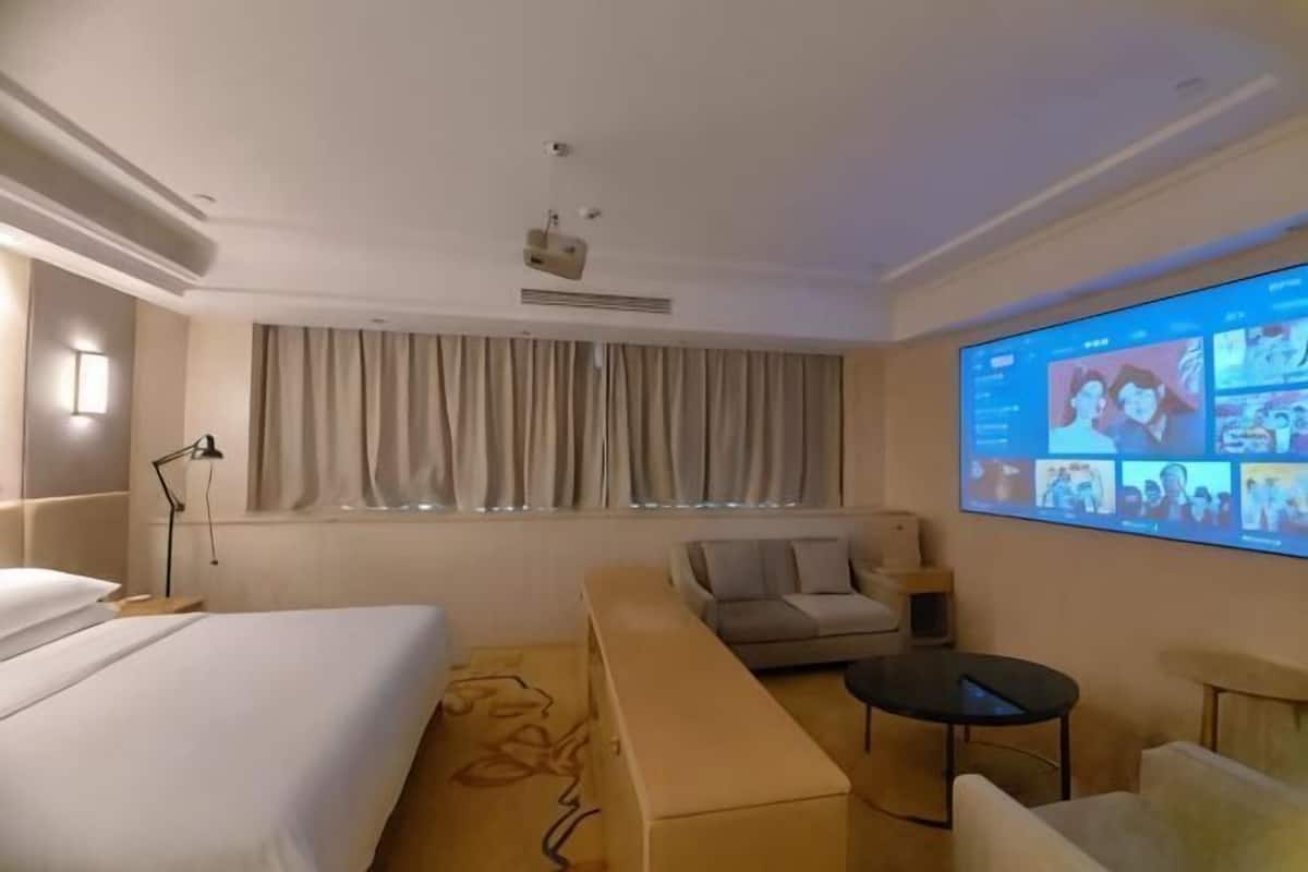 A hotel room with a bed, a sofa, a small table, a projector screen, and a window with curtains.