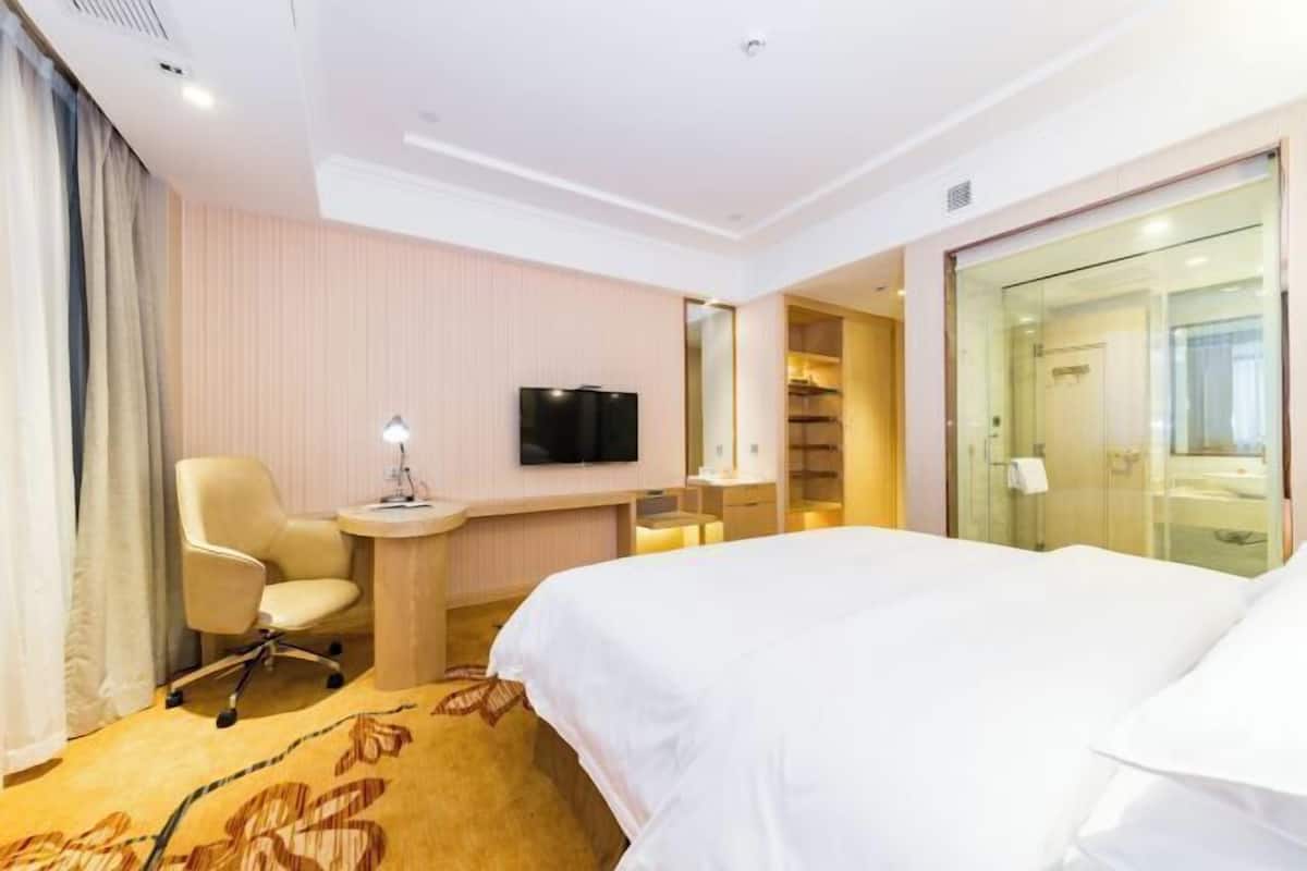 A hotel room with a bed, a desk with a lamp, a TV, a chair, and a glass-enclosed bathroom.