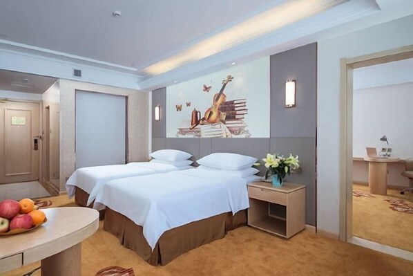 A hotel room with two beds, a desk, a chair, a small table with fruit, and a wall-mounted artwork. - Vienna Hotel (Dongtai Chengdong New District) (Dongtai)