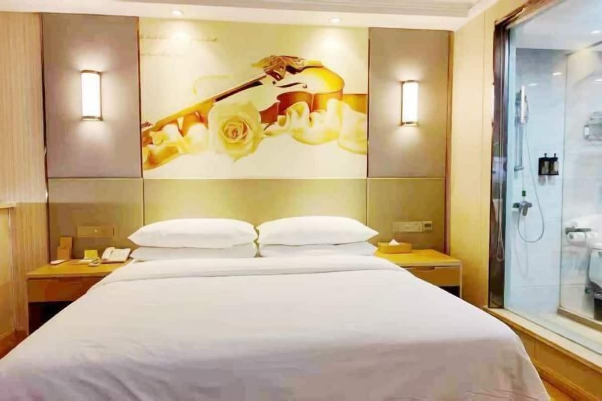 A hotel room with a large bed, two bedside tables, a nightstand, a glass shower enclosure, and a wall-mounted light fixture.