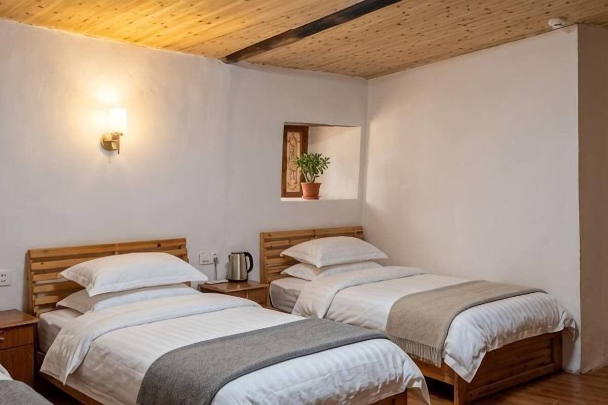 A room with two beds, a wall-mounted light, a small shelf with a plant, and a wooden ceiling.
