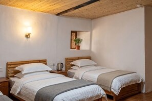 A room with two beds, a wall-mounted light, a small shelf with a plant, and a wooden ceiling.