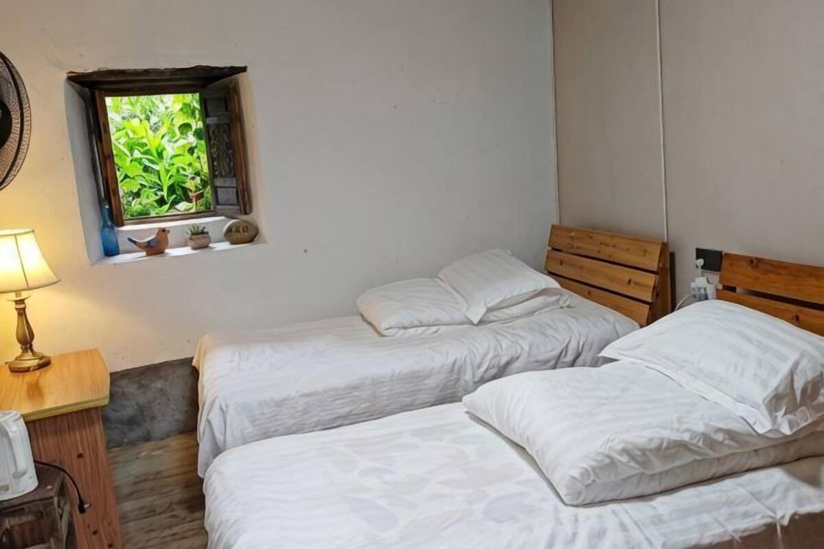 A room with two single beds, a small window with plants, a lamp, and a wooden headboard.