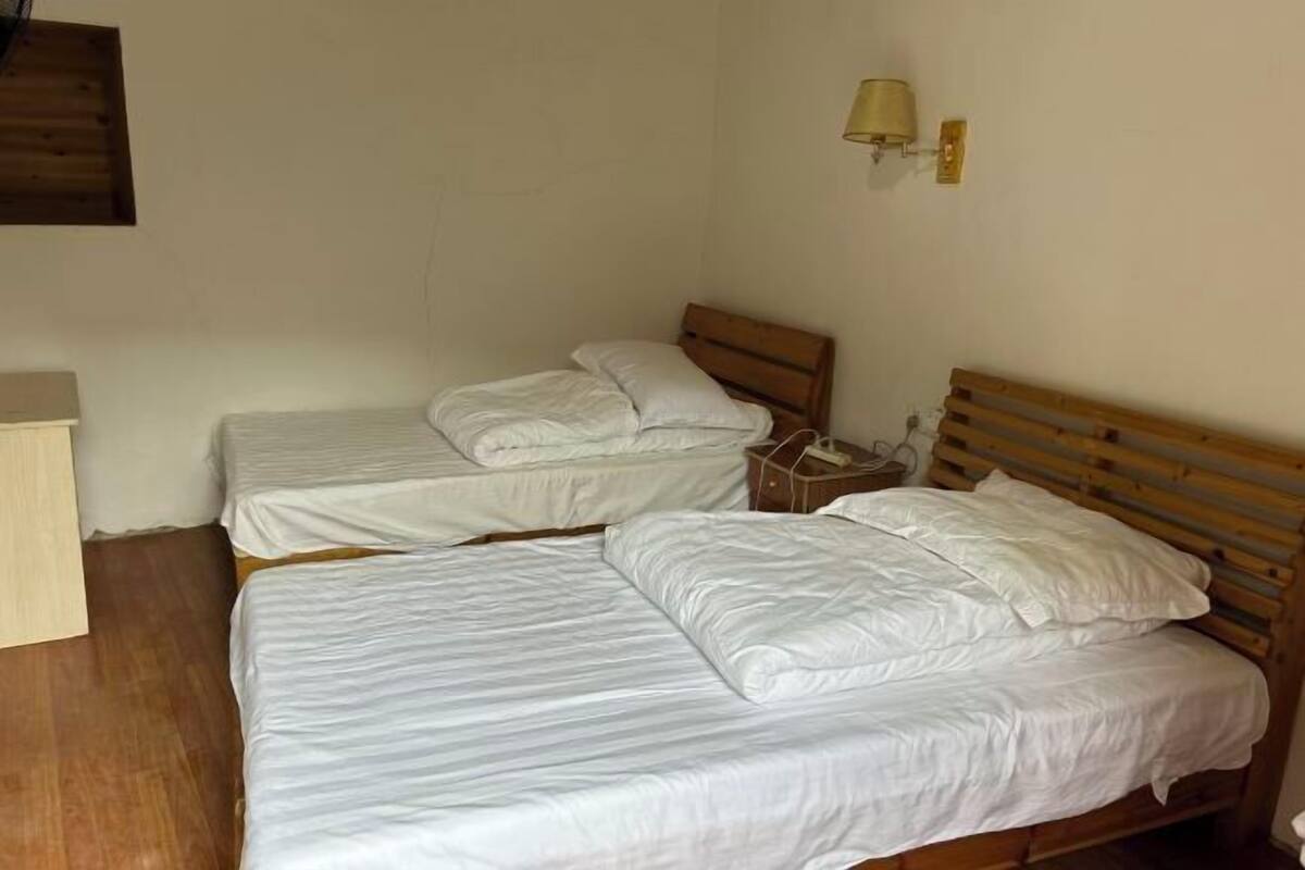 A room with two single beds, wooden headboards, white bedding, a bedside table with a lamp, and a wooden floor.