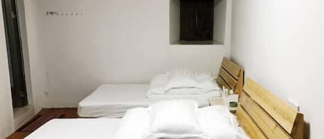 A small, clean room with two single beds, a wooden headboard, a small window, and a door.