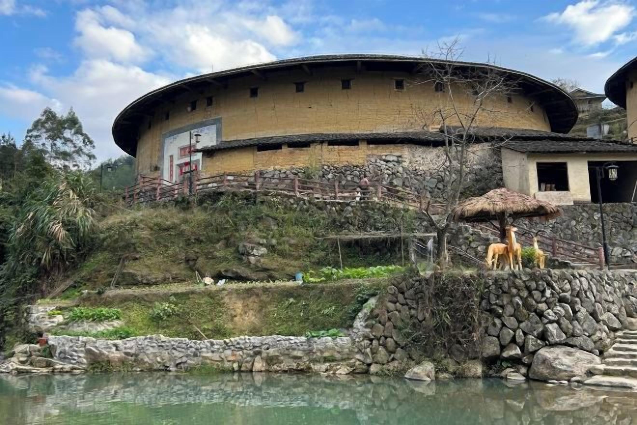 A traditional building with a curved roof, situated beside a body of water, with a stone wall and a thatched-roof structure nearby.
