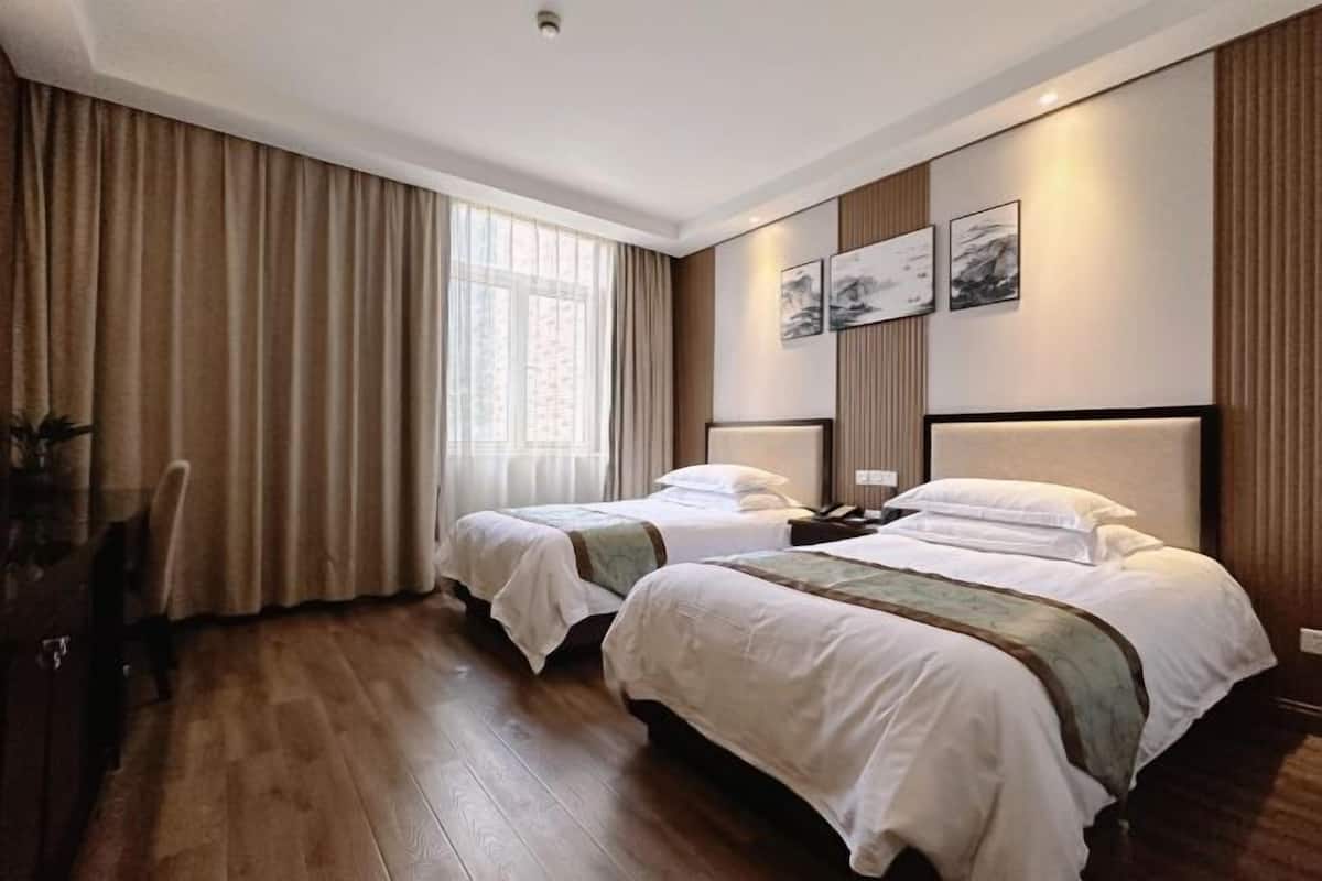 A hotel room with two beds, wooden flooring, a large window with curtains, and artwork on the walls.