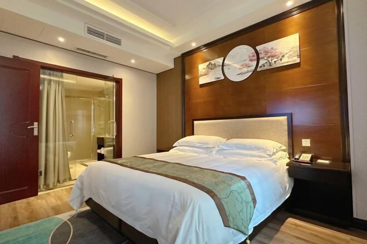 A hotel room with a large bed, a bedside table, a mirror, a wooden headboard, and a wall-mounted clock.
