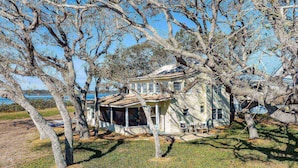 A two-story house with a porch, surrounded by trees and a body of water in the background. - Anchors Away River House (St. Augustine)