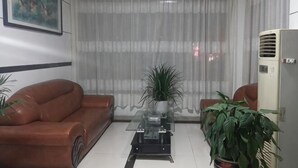 A living room with a brown leather sofa, a glass coffee table, a potted plant, a large window with sheer curtains, and a white air conditioning unit. - Shuangfeng Century Hotel (Shuangfeng)
