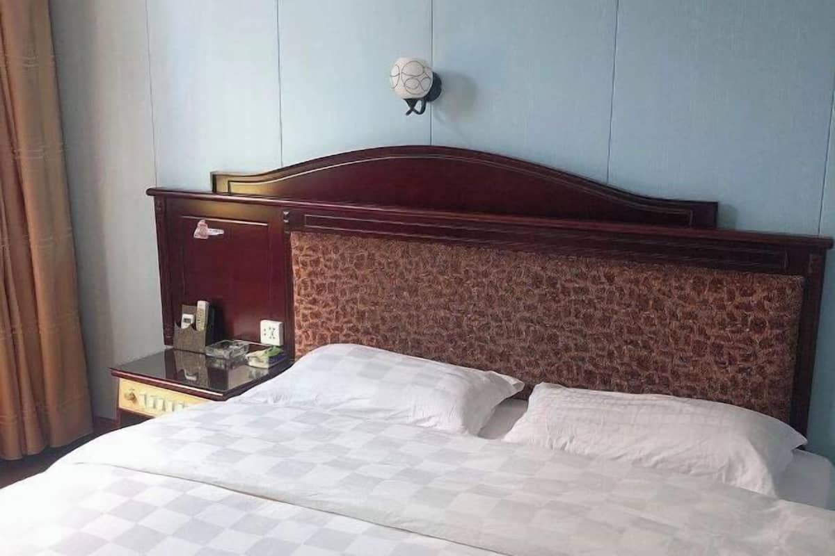 Habitación de hotel con cabecera de madera, cama con ropa blanca, mesita de noche con lámpara y lámpara fijada a la pared.