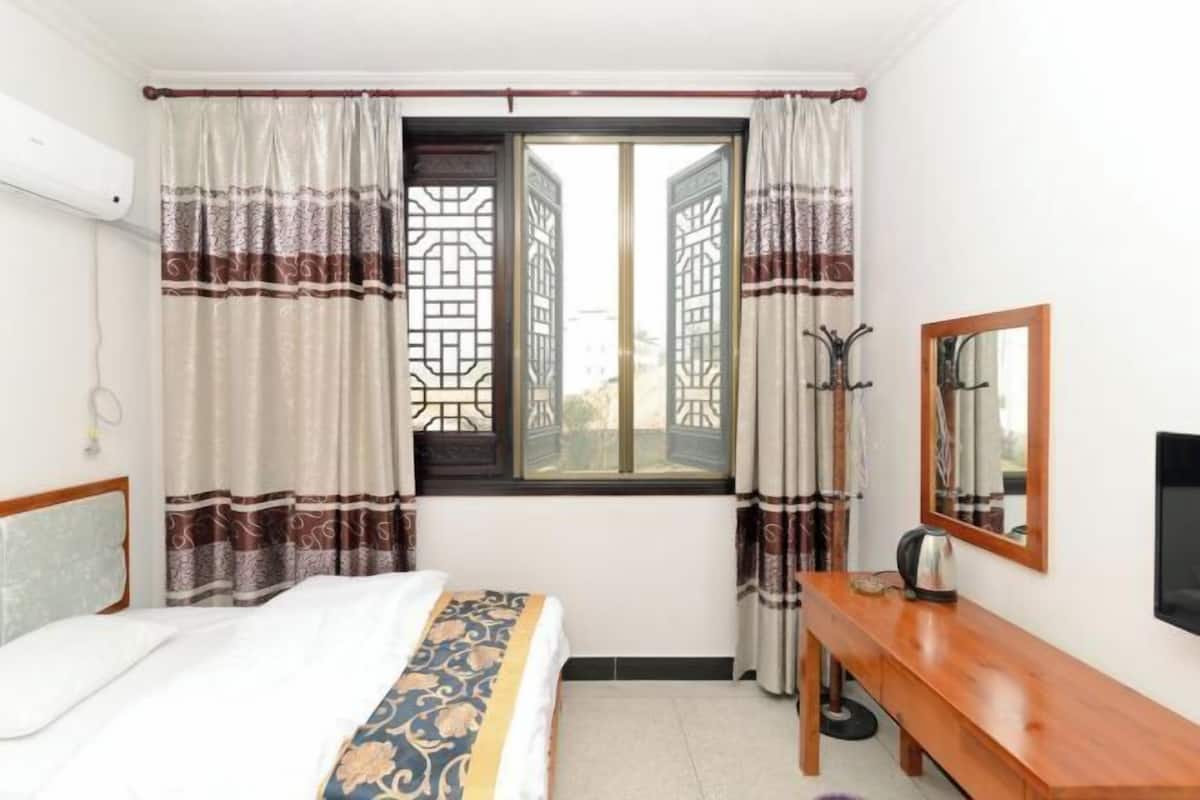 A hotel room with a bed, a wooden bench, a coat rack, a mirror, and a window with decorative curtains.