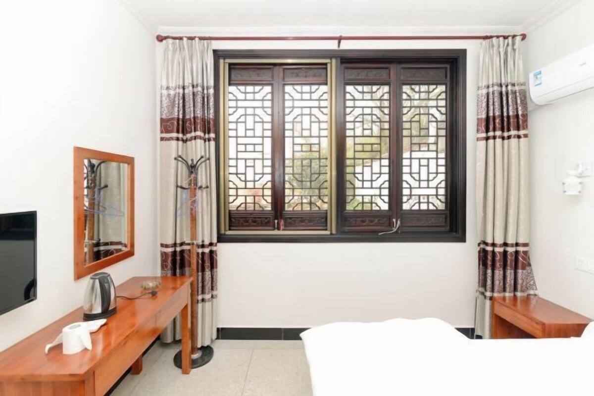 A hotel room with a bed, a wooden desk, a TV, a window with decorative curtains, and an air conditioning unit.