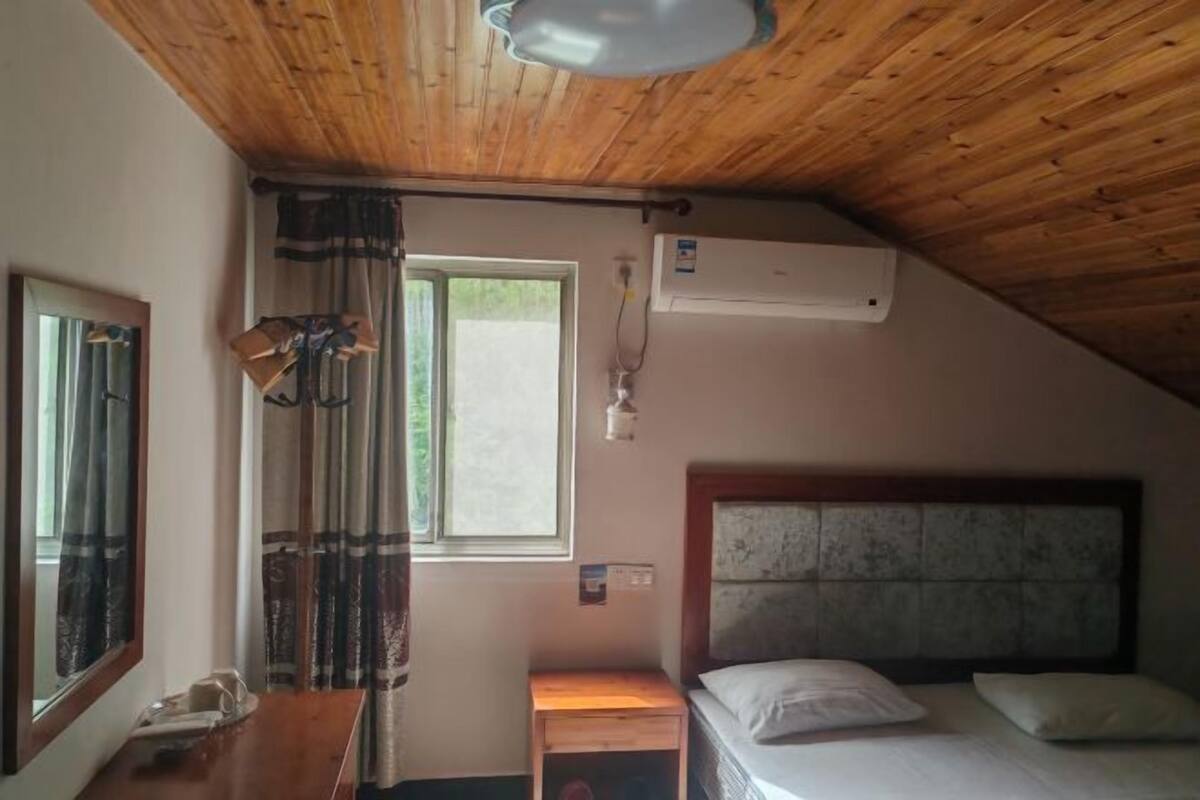 A room with a wooden ceiling, a bed with a headboard, a bedside table, a mirror, a window with curtains, and an air conditioning unit.