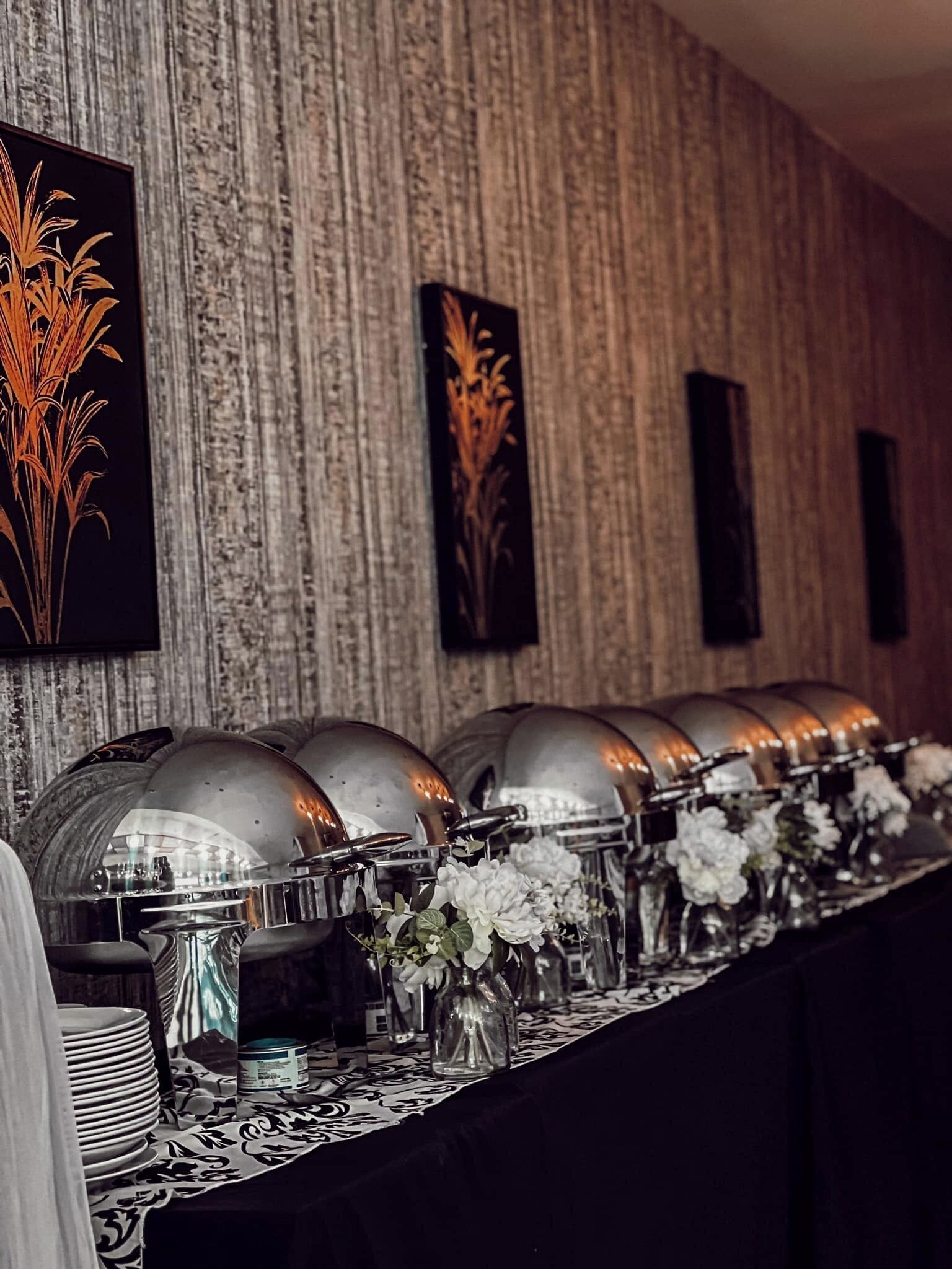 Ein Buffet mit mehreren Speiseauslagen, einer gemusterten Tischdecke und dekorativen Vasen mit Blumen.