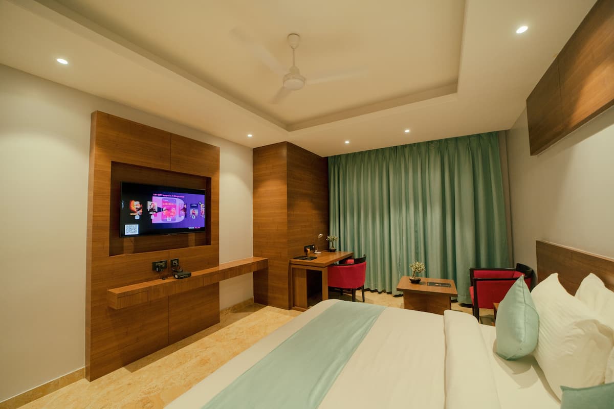 Quarto de hotel com uma cama grande, uma escrivaninha com cadeira, uma televisão na parede e um ventilador de teto.