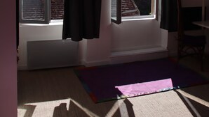 Una habitación con un ventanal grande, un radiador, una colchoneta de yoga morada y una silla.
