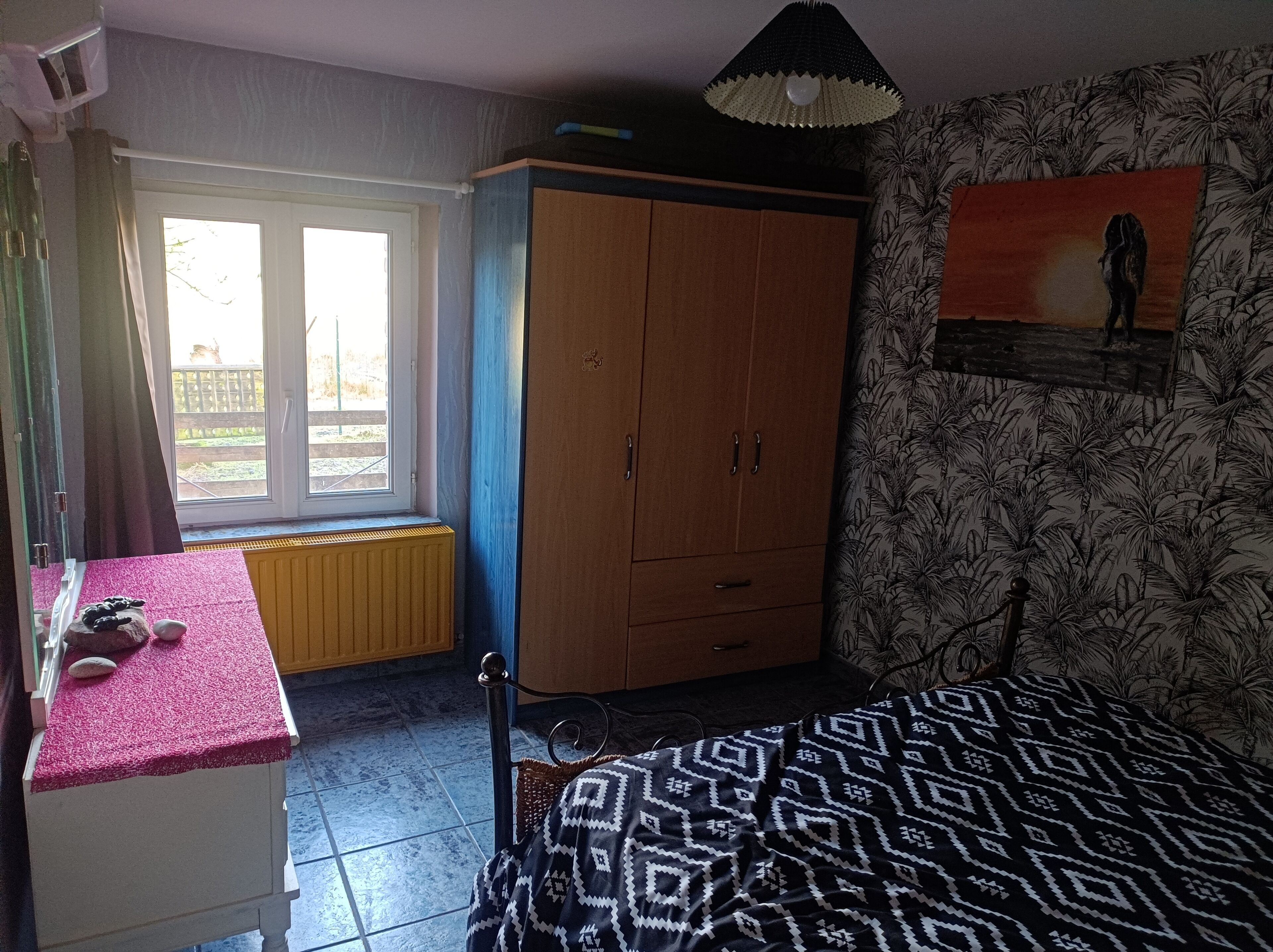 Une chambre à coucher avec un lit, une armoire, un radiateur, une fenêtre et un tableau accroché au mur.