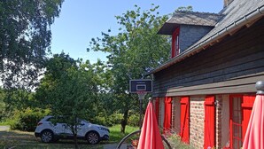 Une terrasse en bois avec des tables de pique-nique et des parasols rouges, un panier de basket et une voiture blanche garée à proximité.