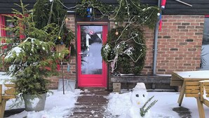 Un sentier enneigé menant à une porte rouge, de part et d’autre de laquelle se trouvent des sapins de Noël en pot et un bonhomme de neige.