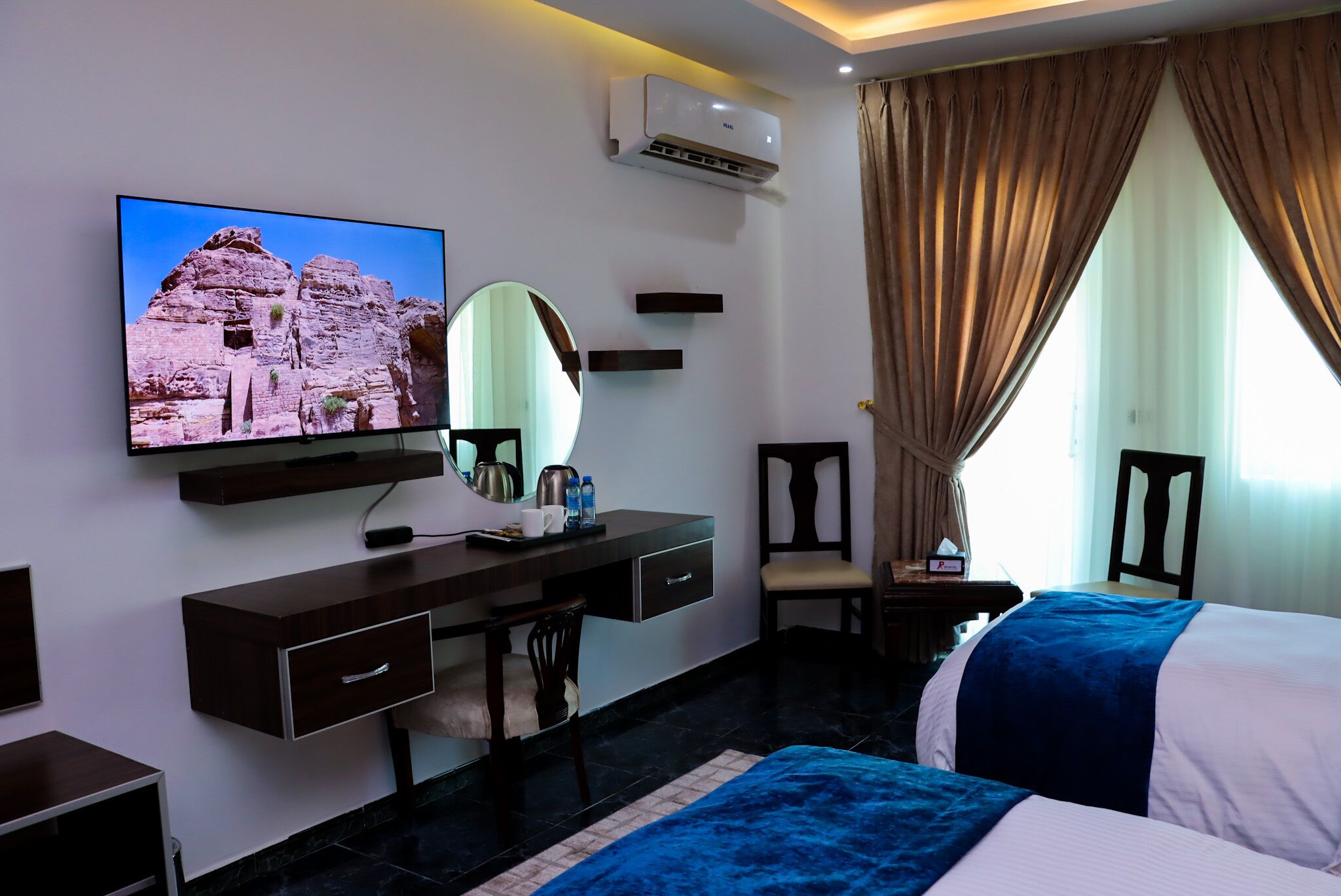 A hotel room with a bed, a desk with a chair, a TV mounted on the wall, a mirror, a window with curtains, and an air conditioning unit.