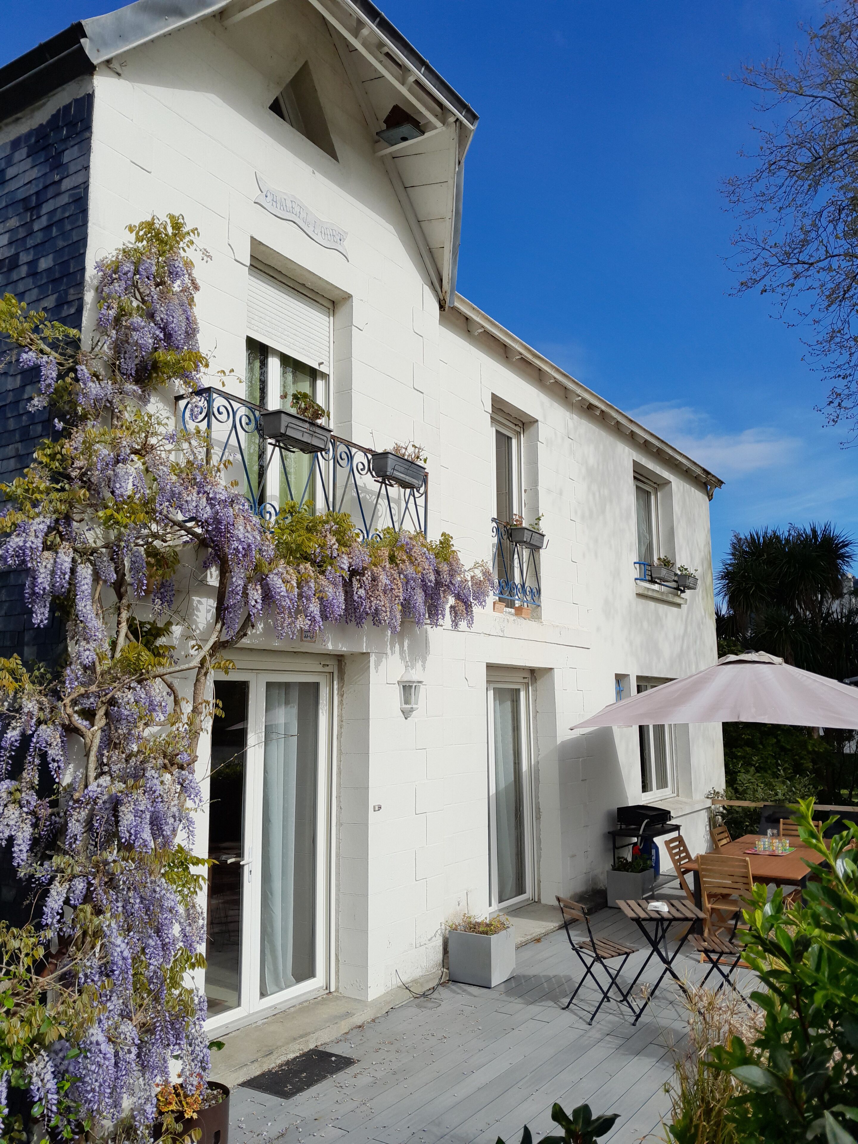 Ein weißes, zweistöckiges Haus mit Balkon, einer Terrasse mit Tisch und Stühlen sowie lila blühenden Weinreben.