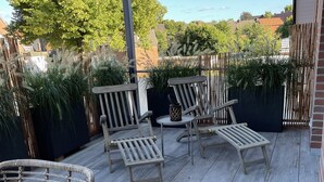 A wooden deck with outdoor furniture, including chairs and a table, surrounded by plants and a glass enclosure. - Exclusive maisonette vacation apartment (Coesfeld)