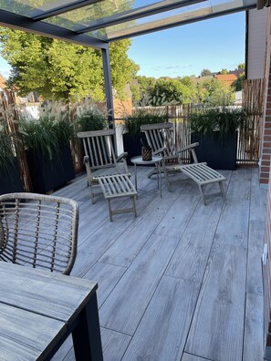 Eine Holzterrasse mit Gartenmöbeln, darunter Stühle und ein Tisch, umgeben von Pflanzen und einer Glaseinfassung.