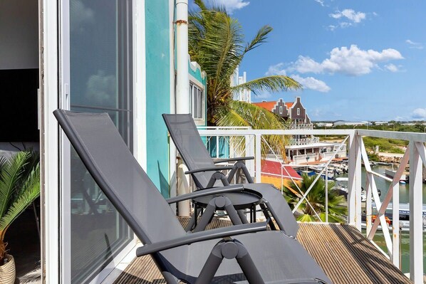 Un balcon avec des fauteuils de jardin, une vue sur un port de plaisance et, au loin, un bâtiment avec un toit rouge.