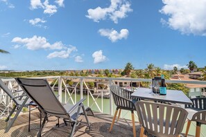 A terrace with a table and chairs, overlooking a body of water and a coastal area with buildings and palm trees.